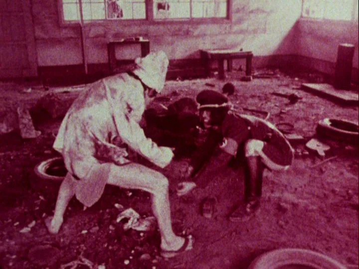Emperor Tomato Ketchup movie still: wide — A dilapidated, trash-strewn space with broken windows and concrete floors. Two individuals crouch on the…