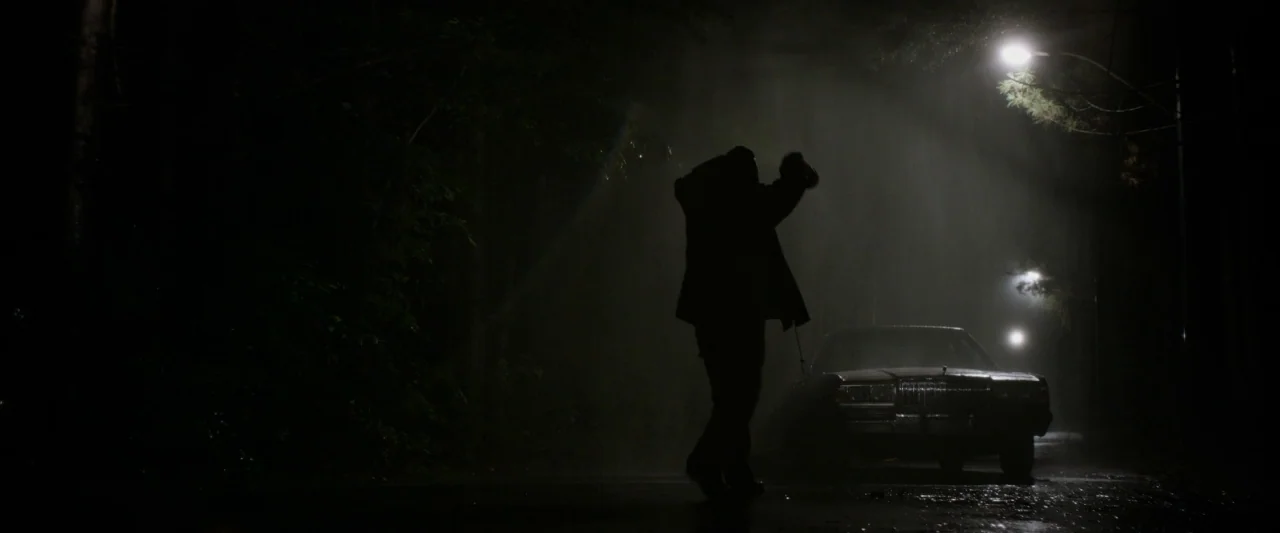 The Monster (2016) movie still: fullbody — A dark, rainy asphalt road at night with dense trees on the left side. A lone figure stands near the…