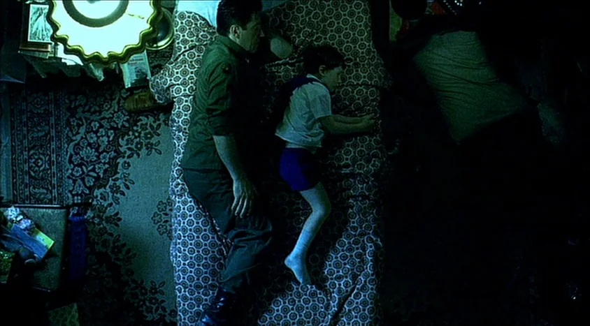 Hedwig and the Angry Inch (2001) movie still: fullbody — A dimly lit room contains a bed with a complex, repeating floral pattern quilt. A man and a…