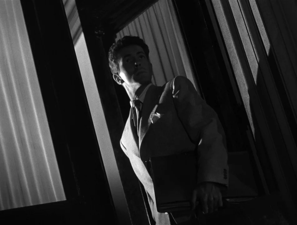 Strangers on a Train (1951) movie still: medium — A man stands framed within a dark doorway illuminated by high-contrast lighting. He holds a…
