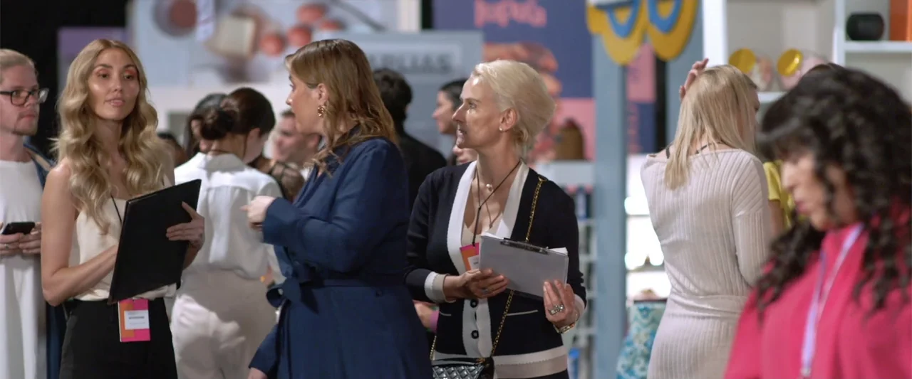Winner Takes the Cake (2025) movie still: medium — An indoor commercial trade show floor features attendees gathered among display booths and…