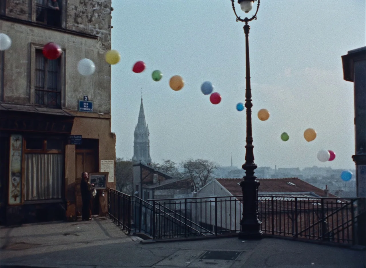 The Red Balloon (1956) movie still: wide — An old Parisian corner features a stone building with a street sign for Rue des Envierges. A string of…