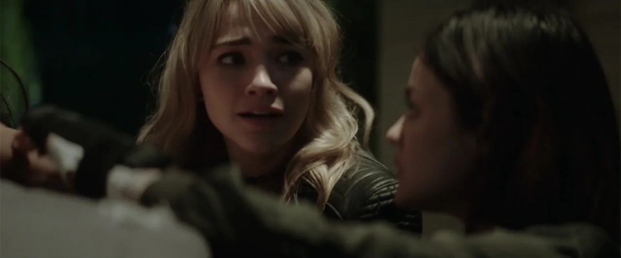 Truth or Dare (2018) movie still: medium — A dark, dimly lit indoor setting focuses on two individuals close together. One blonde woman looks at…