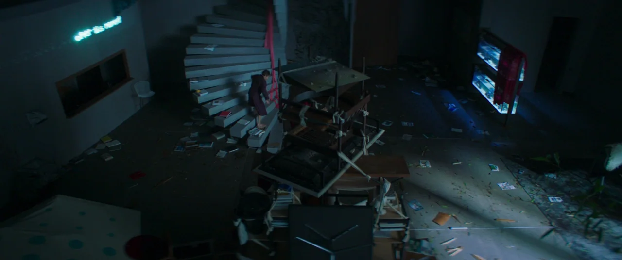 Inside (2023) (2023) movie still: wide — A dark, cluttered living space features a central spiral staircase and scattered papers. A lone girl in a…