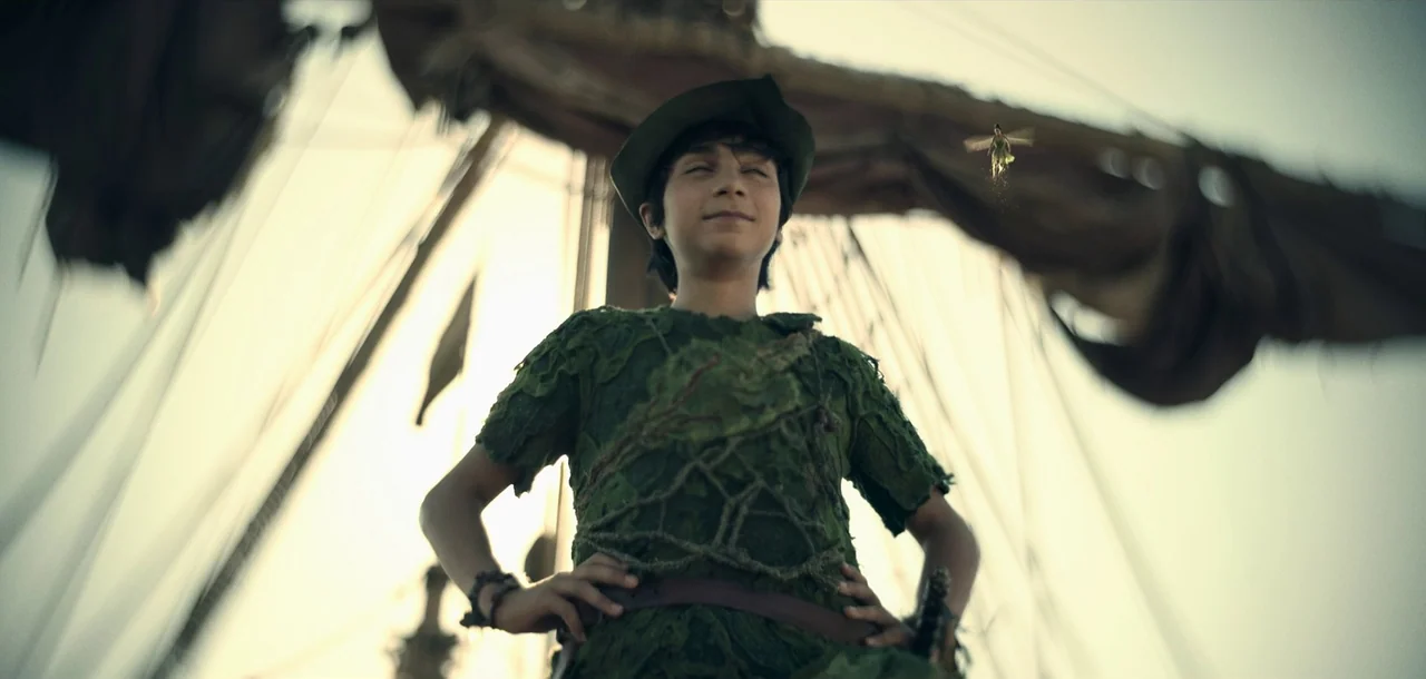 Peter Pan & Wendy (2023) movie still: medium — A boy in a leafy tunic stands on the deck of a wooden sailing ship. He stands with his hands on his…