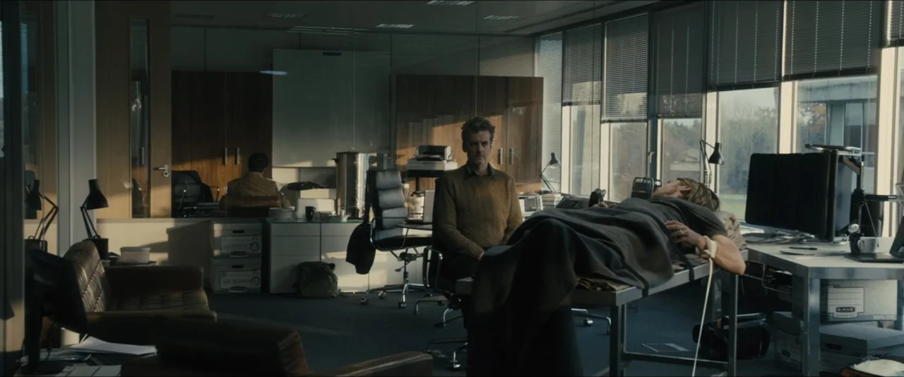 World War Z (2013) movie still: wide — A modern glass-walled office with filing boxes, leather furniture, and computer monitors on desks. A man sits…
