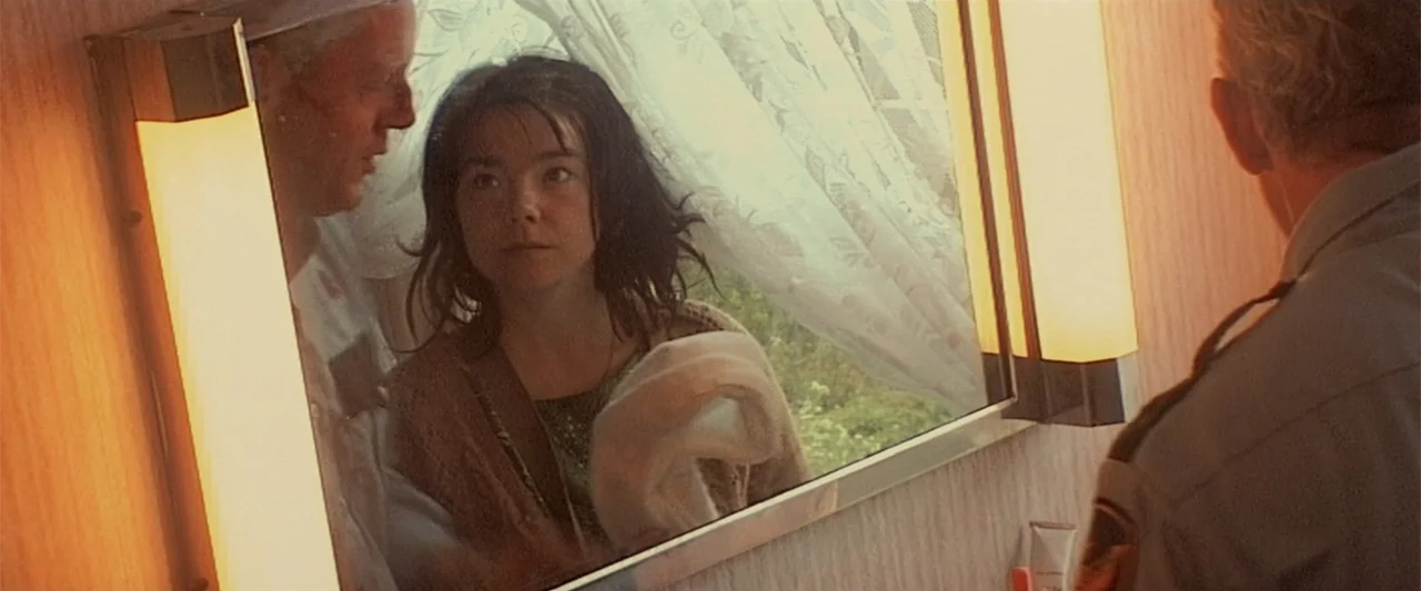 Dancer in the Dark (2000) movie still: medium — A small residential bathroom features a large vanity mirror with flanking light fixtures. A woman…