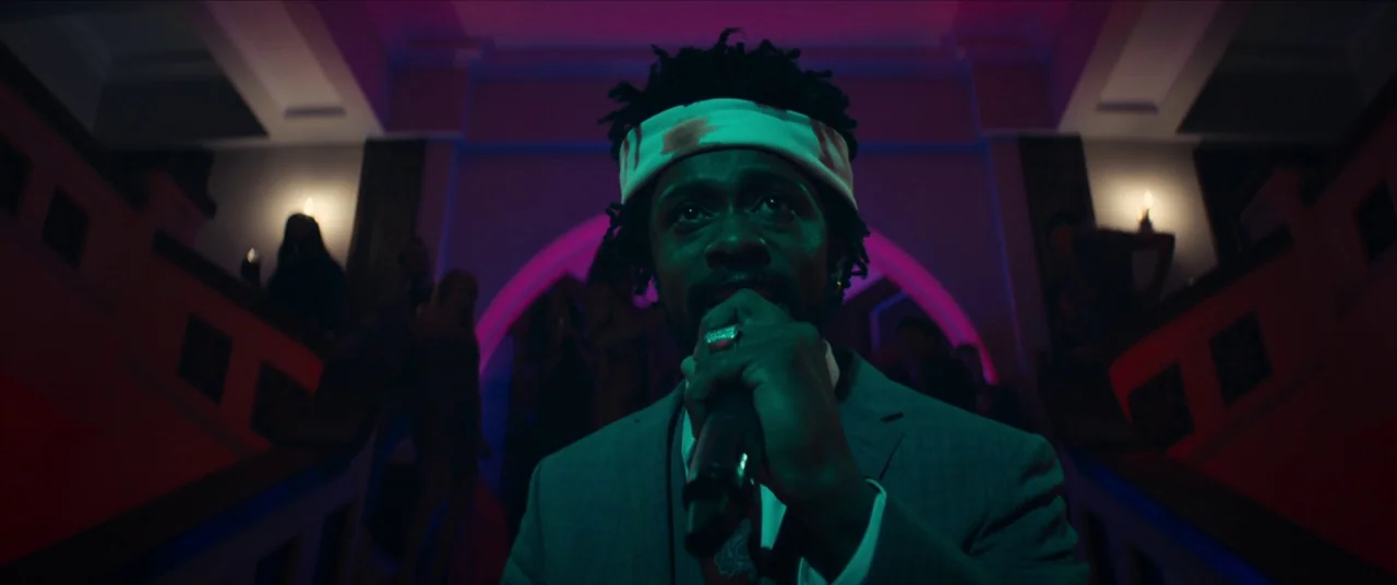 Sorry to Bother You (2018) movie still: medium — A man stands at the base of a grand staircase inside a multi-story building. The man holds a…