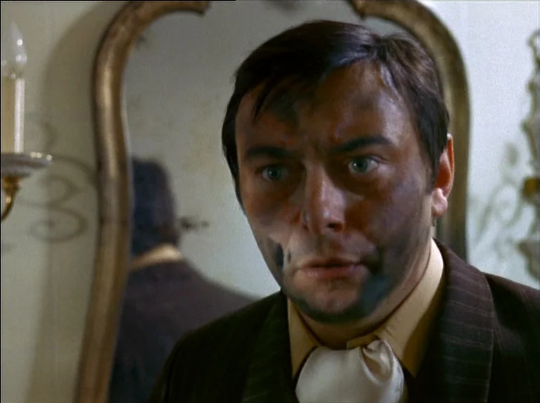 Satan’s Brew (1976) movie still: closeup — A man stands before a large, ornate gilded mirror in a dimly lit room. He has black residue smeared…