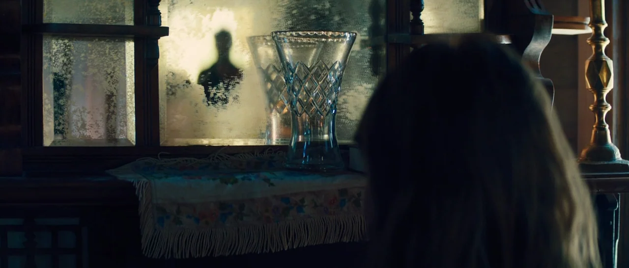 Z for Zachariah (2015) movie still: medium — An antique wooden sideboard with a heavily aged mirror and a decorative lace doily. A person sits out…