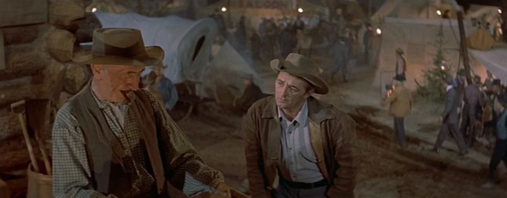 River of No Return (1954) movie still: medium — A bustling mining camp at night features canvas tents and wooden structures under lanterns. Two men…