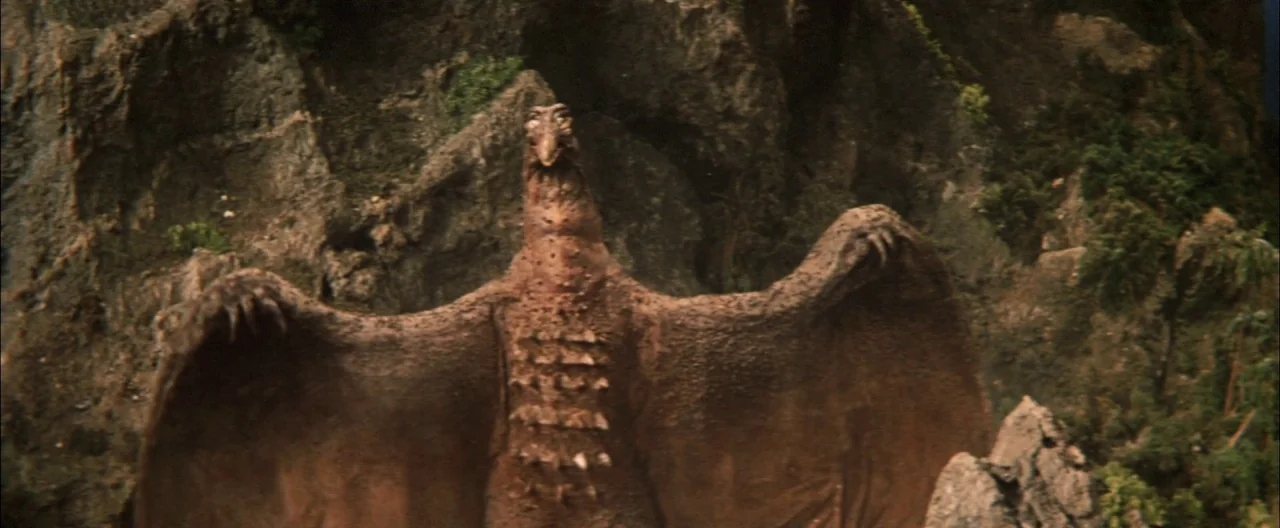 Destroy All Monsters (1968) movie still: fullbody — A craggy rock wall forms a dense natural backdrop behind the creature. The reptilian monster…