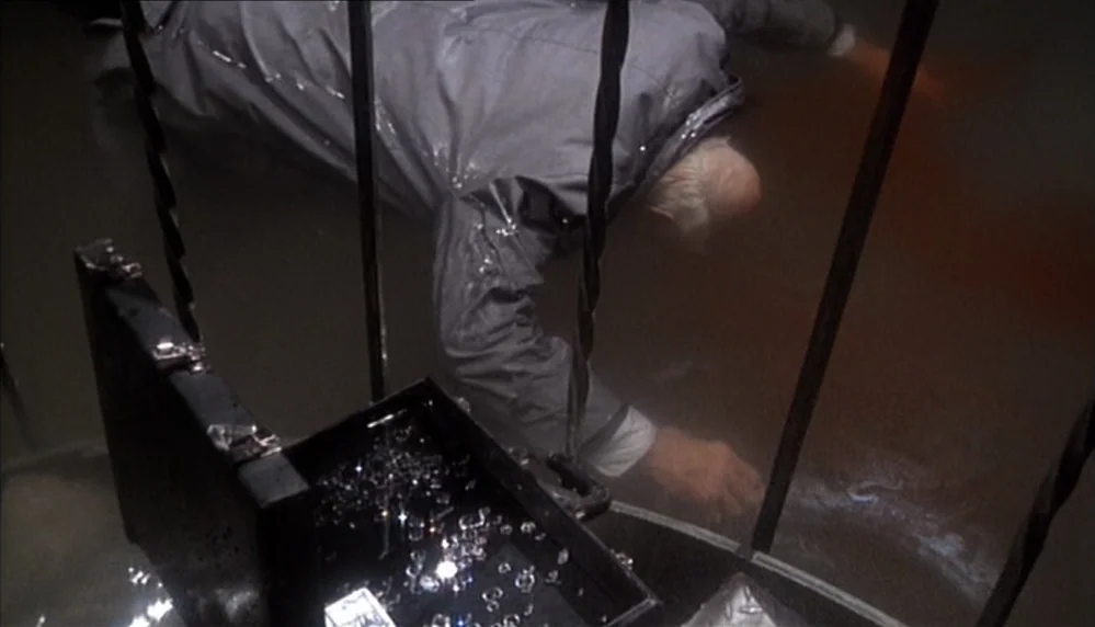 Marathon Man (1976) movie still: medium — A man partially submerged in a dark, flooded space reaches toward a metal case. The open case sits on a…