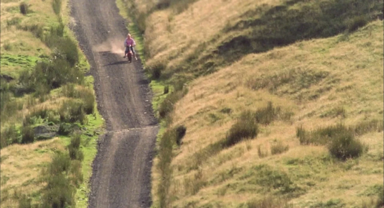 My Summer of Love movie still: wide — A remote dirt trail cuts through a vast, grassy hillside with sparse vegetation. A man rides a small red…