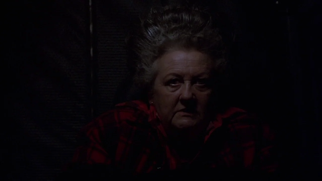 Pee-Wee’s Big Adventure (1985) movie still: closeup — A dimly lit interior space with a dark, indistinct background. An older woman in a red plaid…