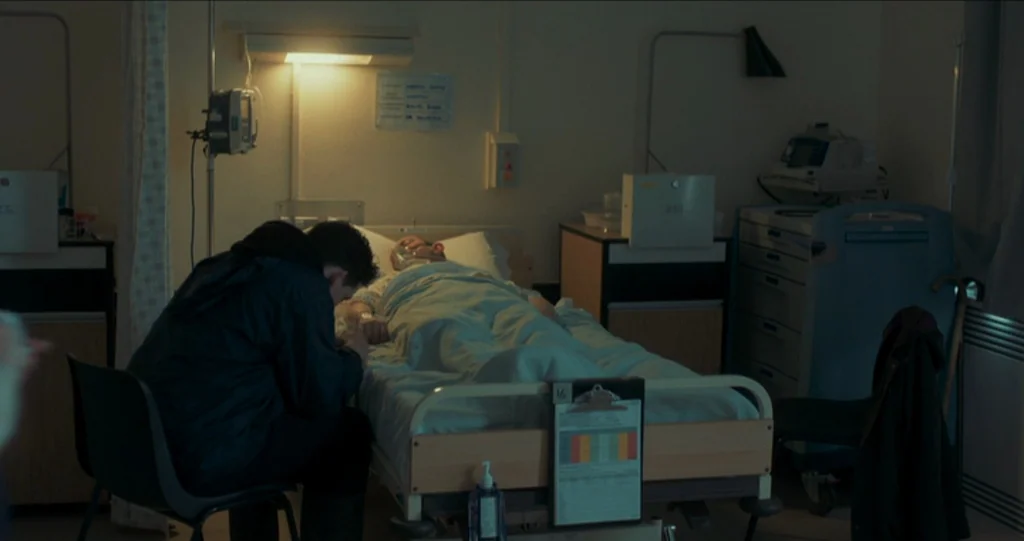 God’s Own Country (2017) movie still: medium — A quiet hospital room contains a patient in bed and a visitor seated beside him. The visitor leans…