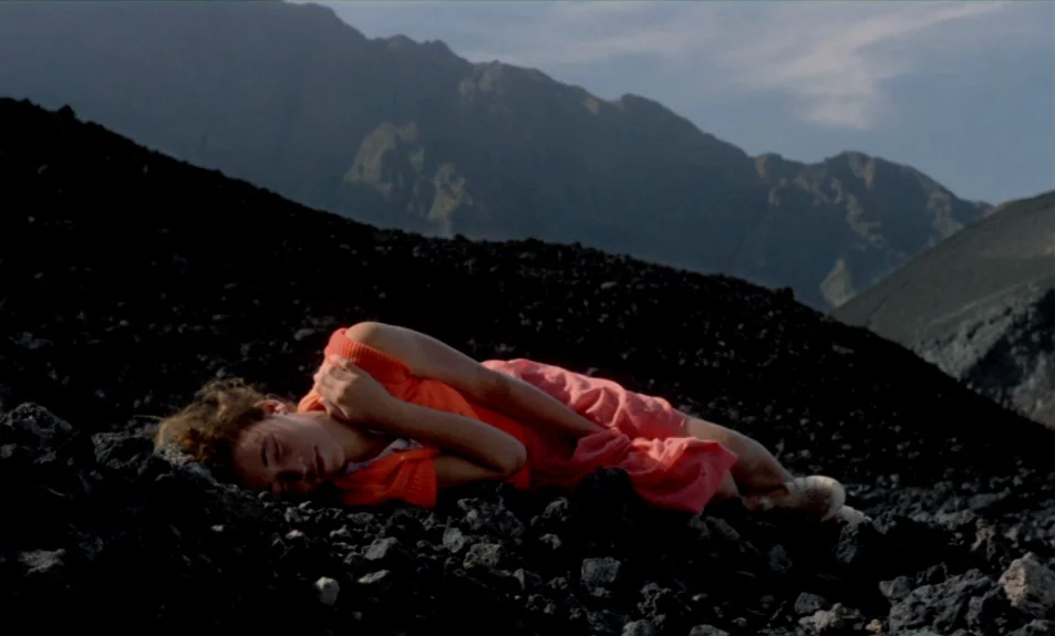 Casa De Lava (1994) movie still: medium — A desolate volcanic landscape with steep dark mounds and distant layered mountain ridges. A woman lies…