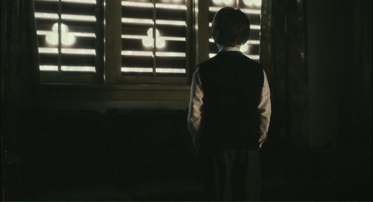 El hijo del diablo (1966) movie still: medium — A dimly lit room with heavy curtains flanking a large window. A young boy stands facing the window…