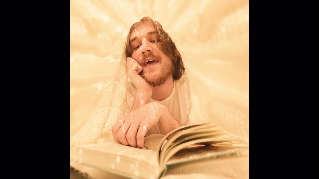 Bo Burnham: Inside (2021) movie still: closeup — A man lies under a white textured fabric blanket draped like a canopy. He rests his chin on his…