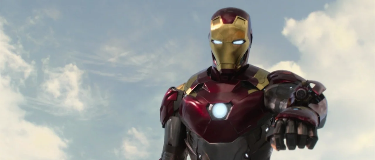 Spider Man: Homecoming (2017) movie still: medium — A high-tech armored figure stands against a bright, cloudy daytime sky. The character extends…