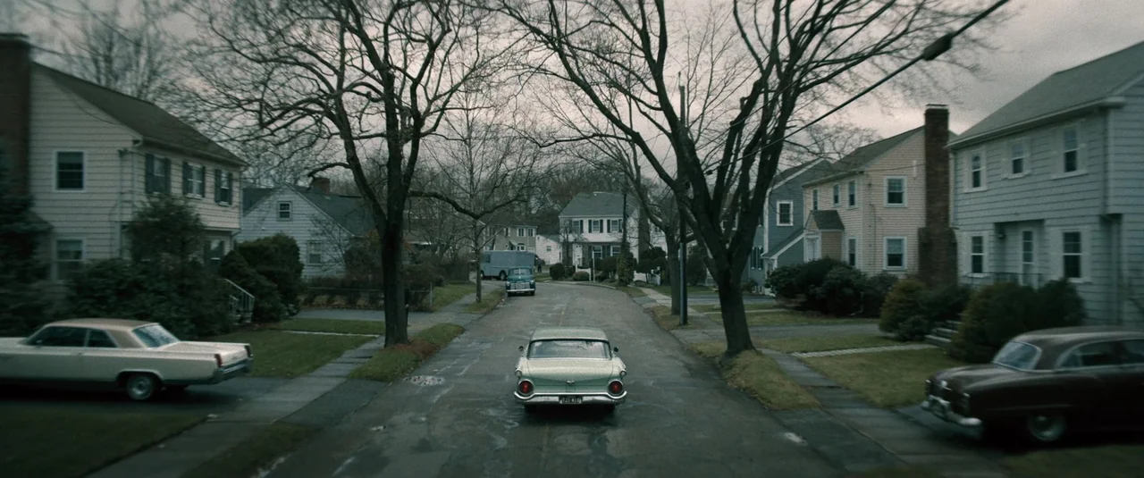 Boston Strangler (2023) movie still: wide — A quiet suburban street lined with two-story houses and bare leafless deciduous trees. A vintage car…