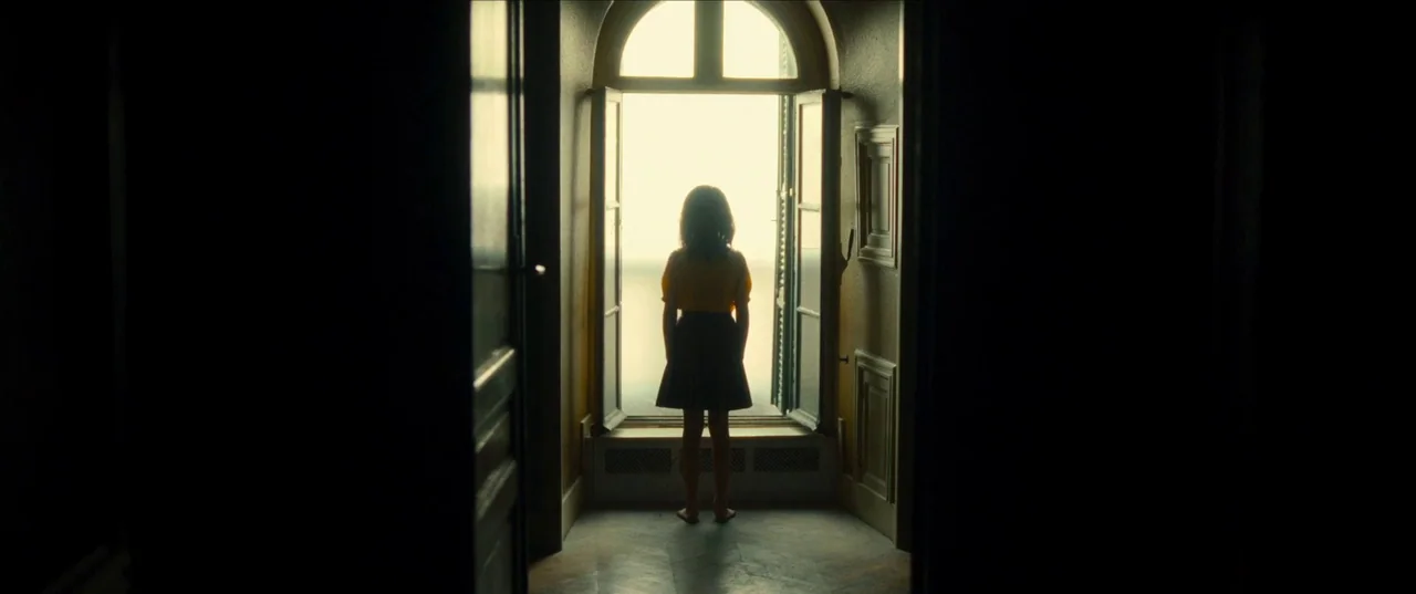 Earwig (2022) movie still: fullbody — A dark hallway leads to a brightly lit open window overlooking a white space. A young girl in a yellow shirt…