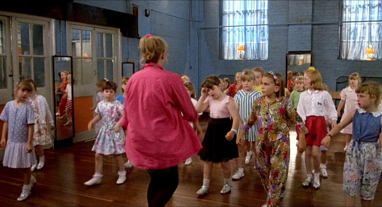 Life Is Sweet (1990) movie still: medium — A group of young girls in colorful patterned dresses and outfits gather in a studio. The instructor in a…