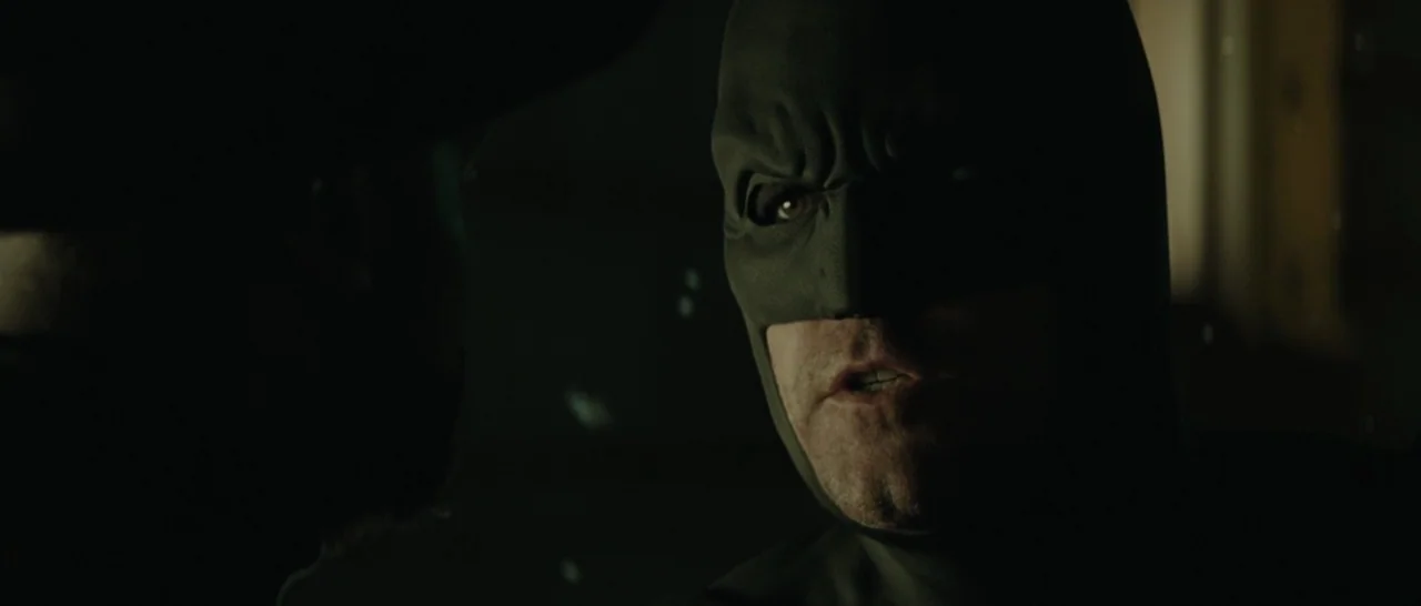 Suicide Squad (2016) movie still: closeup — A dimly lit cabin of a high-tech vehicle with dark surfaces. A man wearing a grey rubber mask speaks…