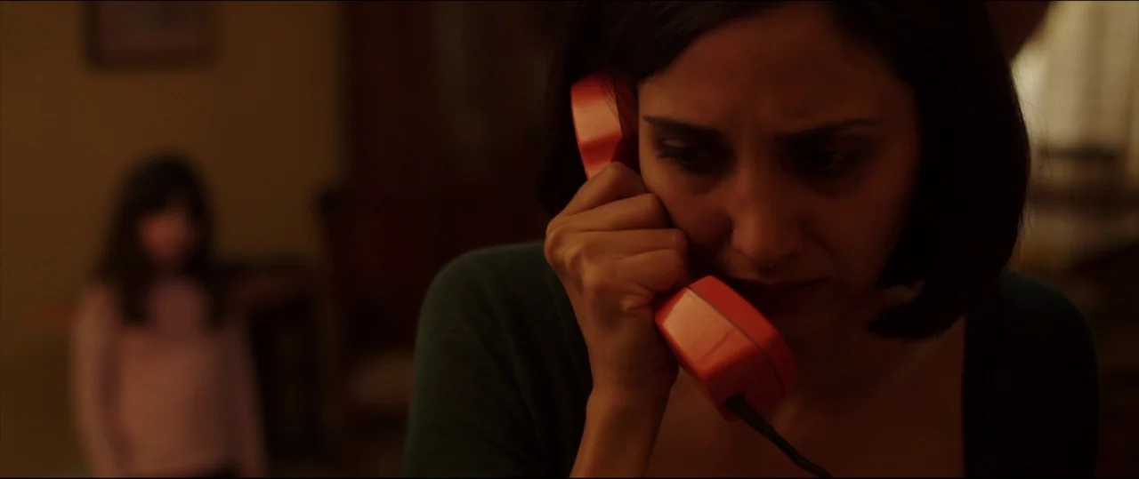 Under The Shadow (2016) movie still: closeup — A dark, dimly lit domestic space with a woman in the immediate foreground. A young girl stands out of…
