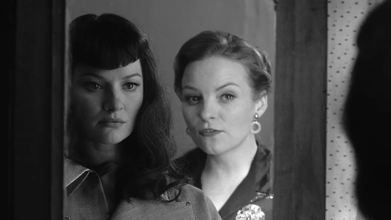 The Notorious Bettie Page (2005) movie still: closeup — Two women view their reflections in a mirror within a domestic space. The foreground woman…