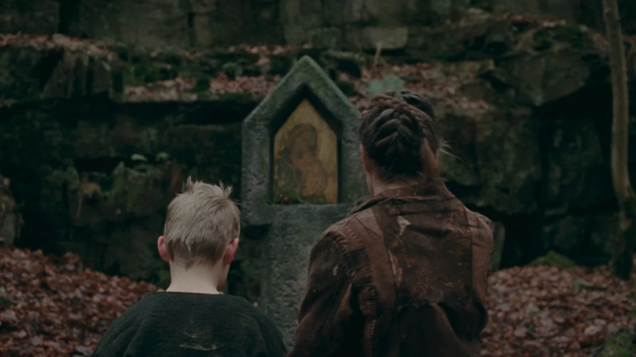 The Devil’s Bath (2024) movie still: medium — A stone religious shrine sits nestled against a mossy rock face in a forest. A woman and a young boy…