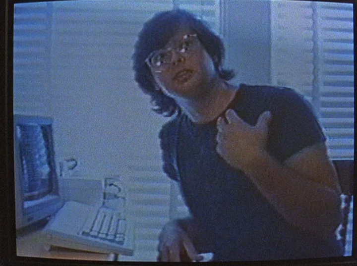 Slacker (1991) movie still: medium — A dimly lit home office space features window blinds and a vintage computer monitor. A young person in a dark…