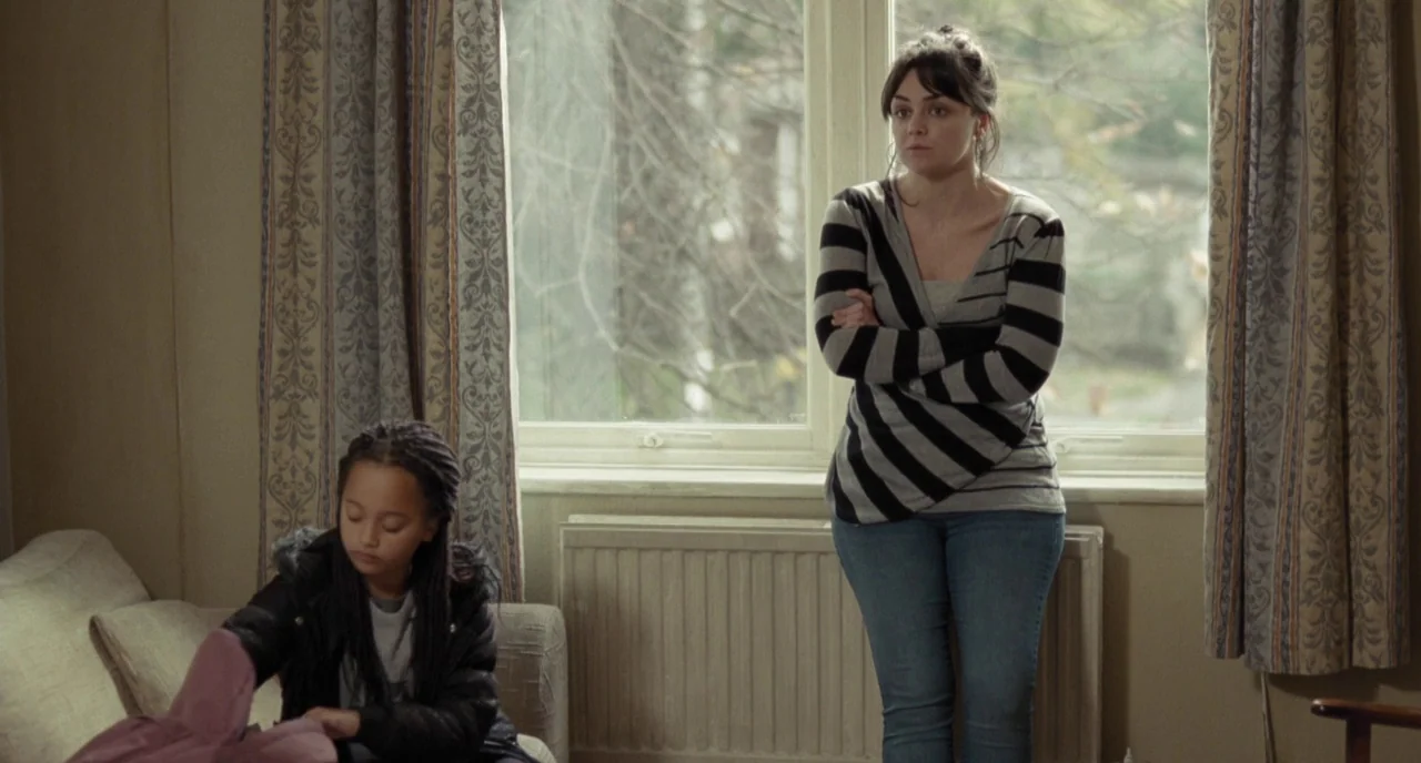 I, Daniel Blake (2016) movie still: medium — A residential room features a window with patterned curtains and a white radiator below. An adult woman…