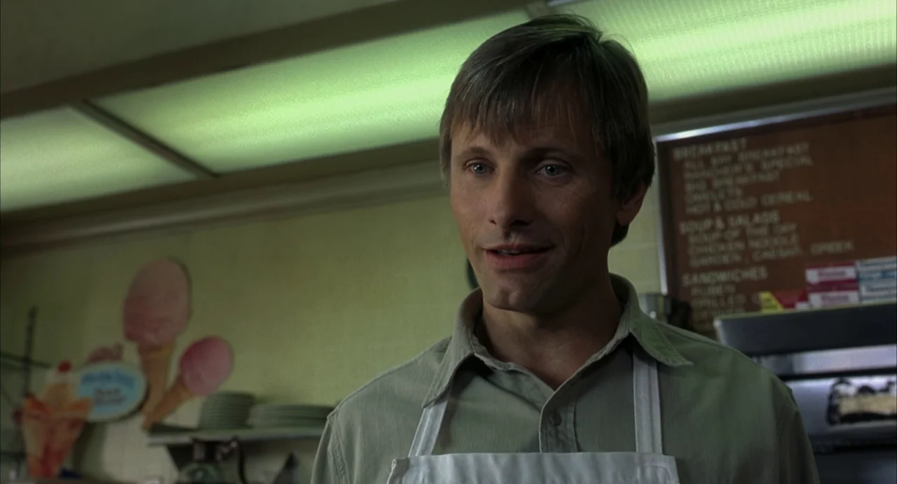 A History of Violence (2005) movie still: medium — The interior of a diner features a menu board and ice cream cone wall art. A man wearing an apron…
