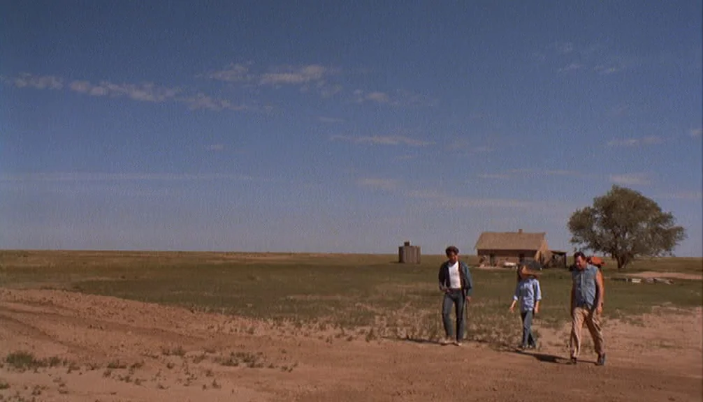 Badlands (1973) movie still: wide — A flat, sparsely vegetated plain stretches toward a small house and a single tree. Three people walk across the…