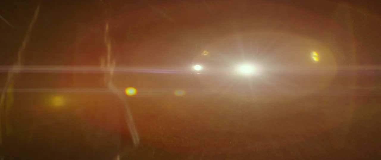 The Strangers: Prey at Night (2018) movie still: wide — A dark, unlit road at night with tall grass lining the edges. A vehicle approaches from the…