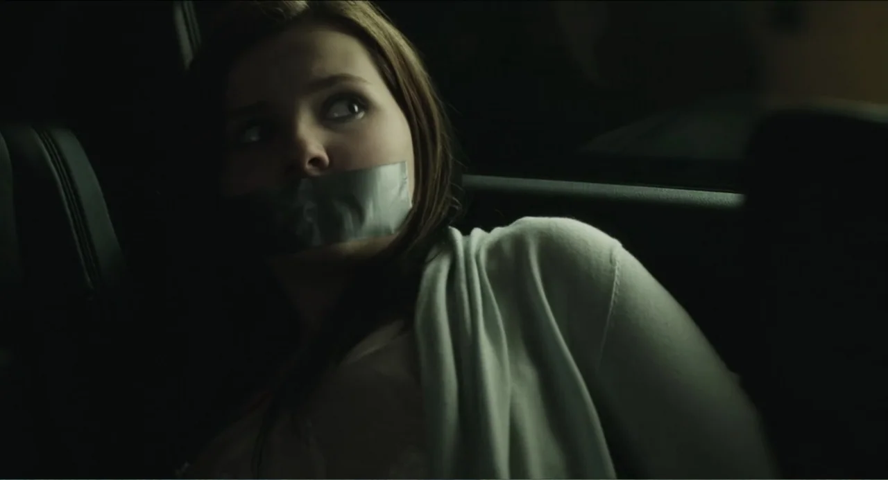 Haunter (2013) movie still: closeup — The interior of a dark car is visible at night with faint light entering. A young woman with long hair and a…