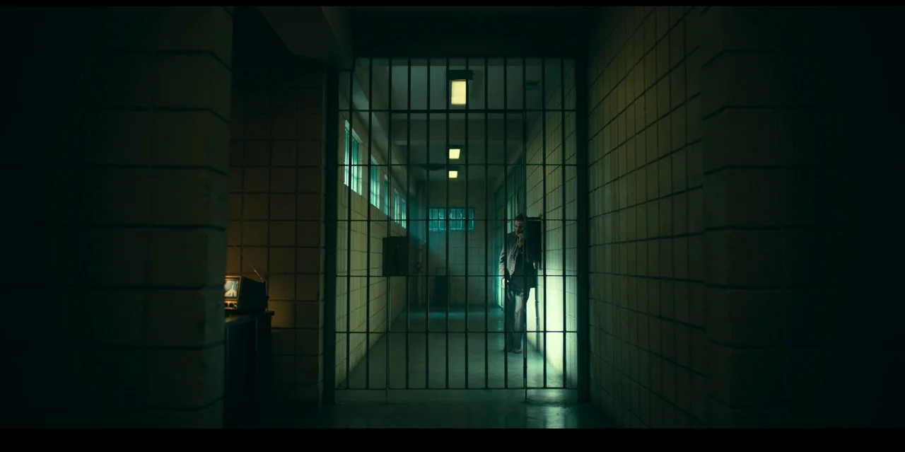 Joker: Folie à Deux (2024) movie still: wide — A dark hallway with beige cinder block walls is blocked by a heavy metal gate. A man in a suit stands…