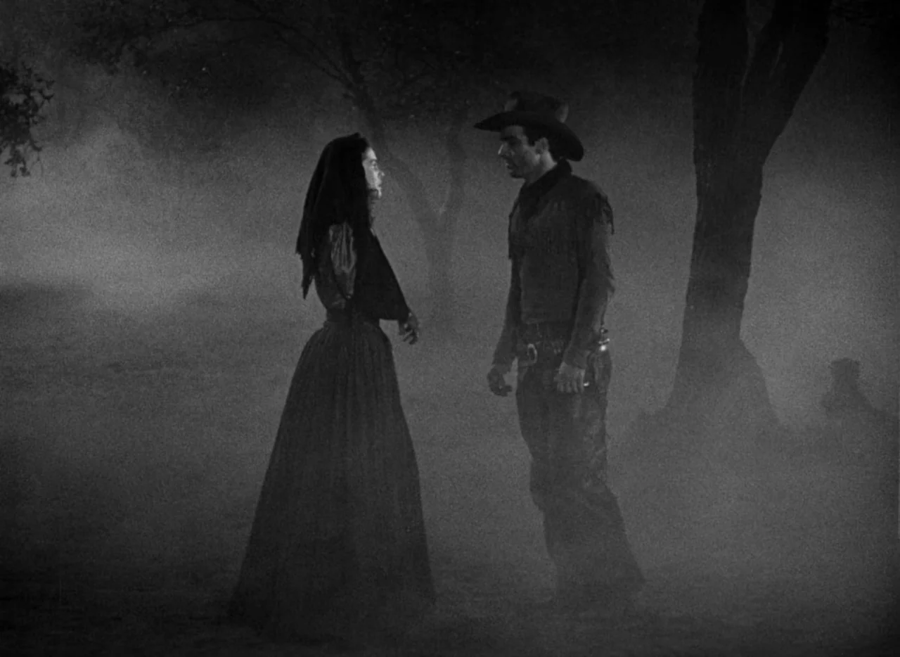 Red River (1948) movie still: fullbody — A dense, foggy outdoor area features dark trees looming in the background. A woman in a long dress and a…