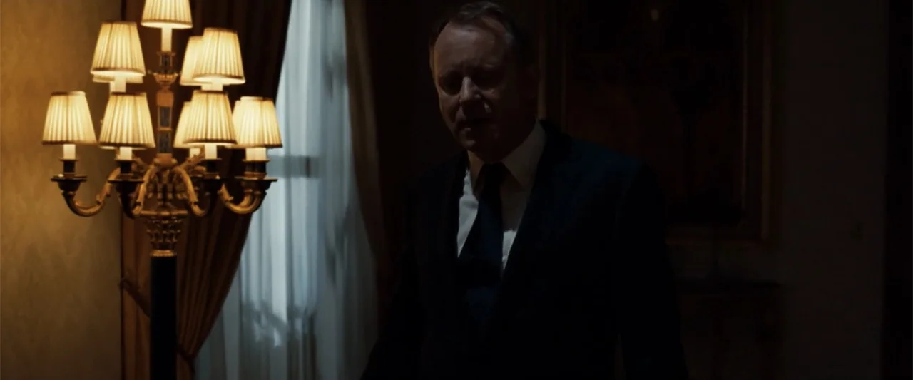 Angels & Demons (2009) movie still: medium — An opulent room with heavy curtains and a gilded floor lamp at night. A middle-aged man in a dark suit…