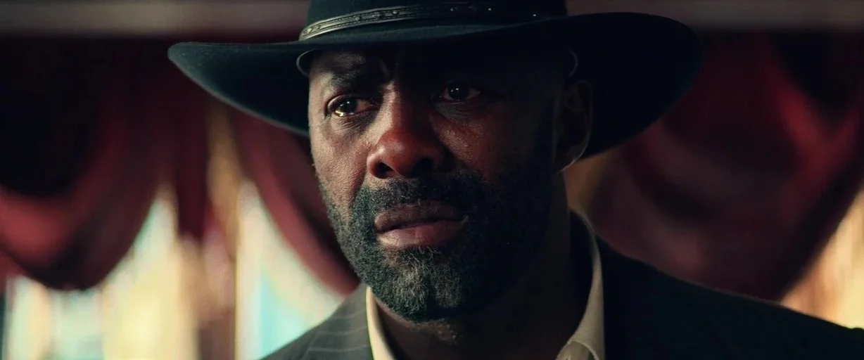 The Harder They Fall (2021) movie still: closeup — A man stands before draped red curtains in a dimly lit indoor space. He is looking ahead with an…