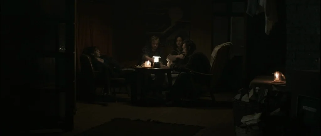 One Hundred Mornings (2009) movie still: wide — A dimly lit room with wooden walls is occupied by four people at night. They sit around a small…