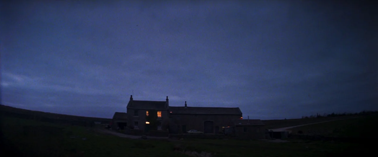 Starve Acre (2023) movie still: establishing — A stone farmhouse stands isolated under a vast, dark, cloudy twilight sky. Yellow light beams from…