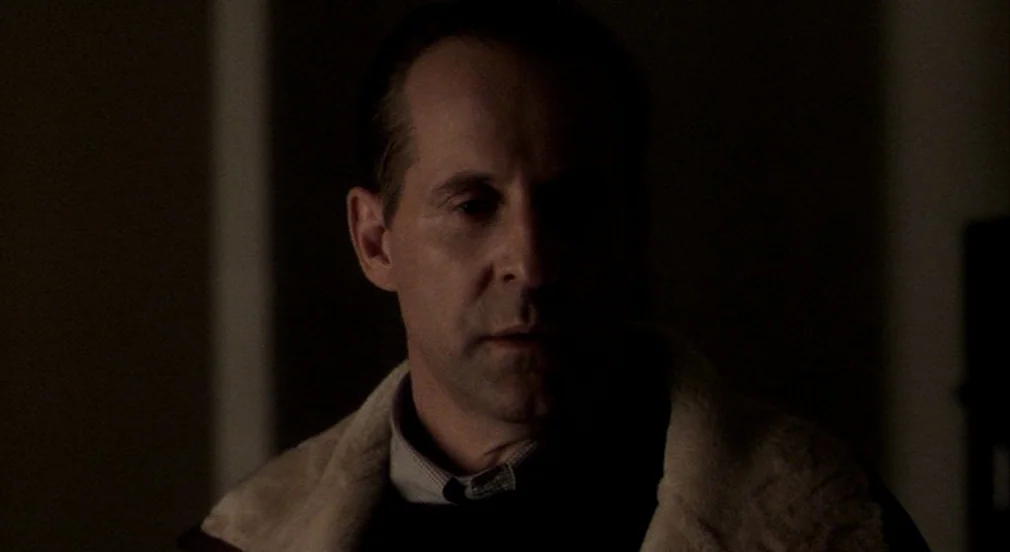 Birth (2004) movie still: closeup — A dimly lit interior space with a dark, indistinct background. A middle-aged man gazes toward the left side of…
