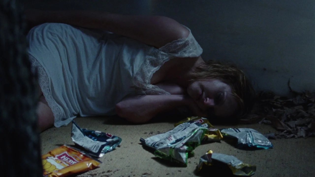 Queen of Earth (2015) movie still: medium — A dim room has a woman lying on a dark carpeted floor. Several crumpled snack bags are scattered around…