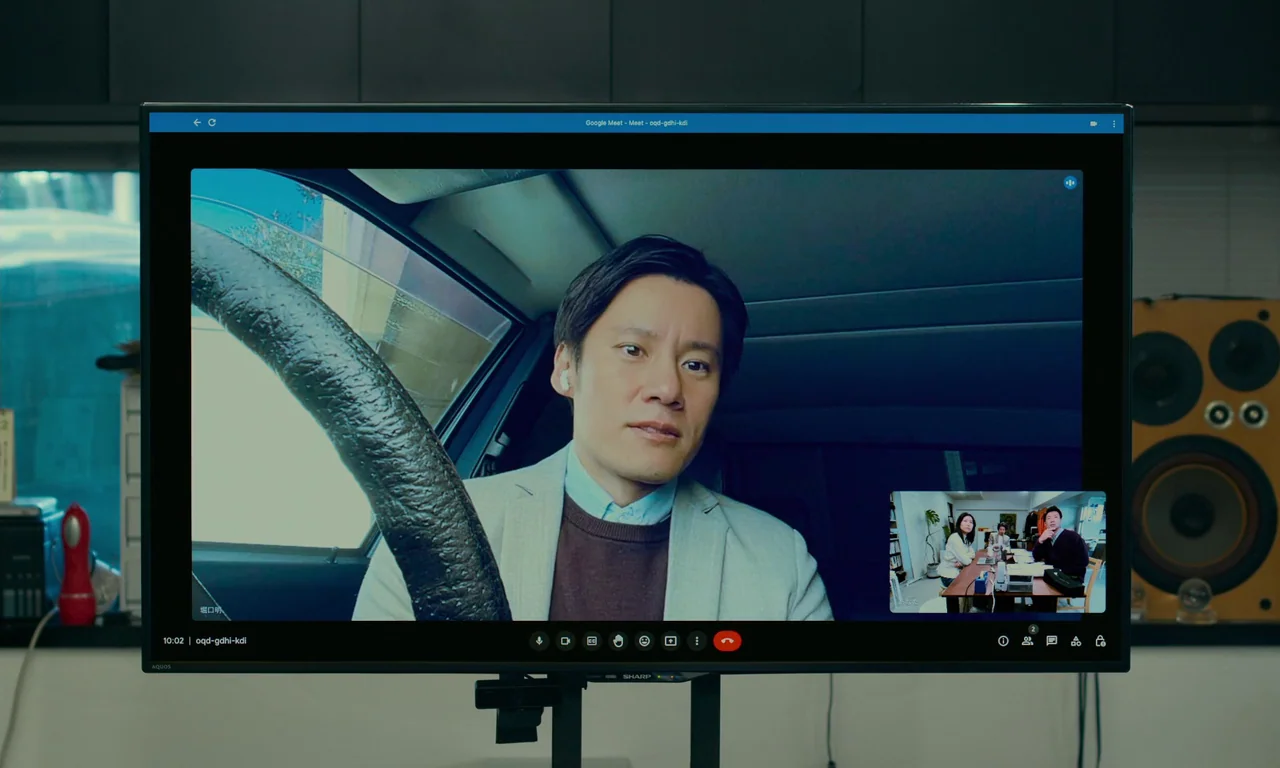 Evil Does Not Exist (2023) movie still: medium — A Sharp television screen displays a video conference call featuring a man in a car. The man…