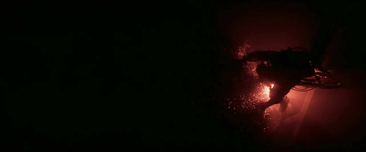 Last Breath (2025) movie still: wide — A dark corridor features a single bright red light source on the floor. A person in dark clothing crouches…