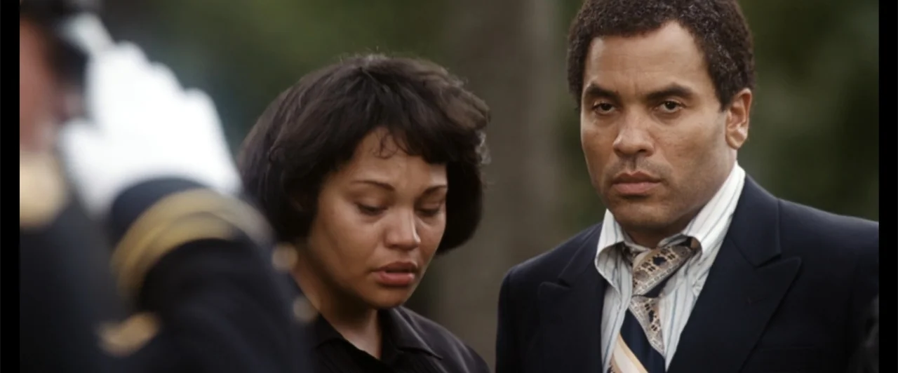 Exposed (2016) movie still: medium — A man and woman stand together outdoors against an out-of-focus background. They appear to be attending a…