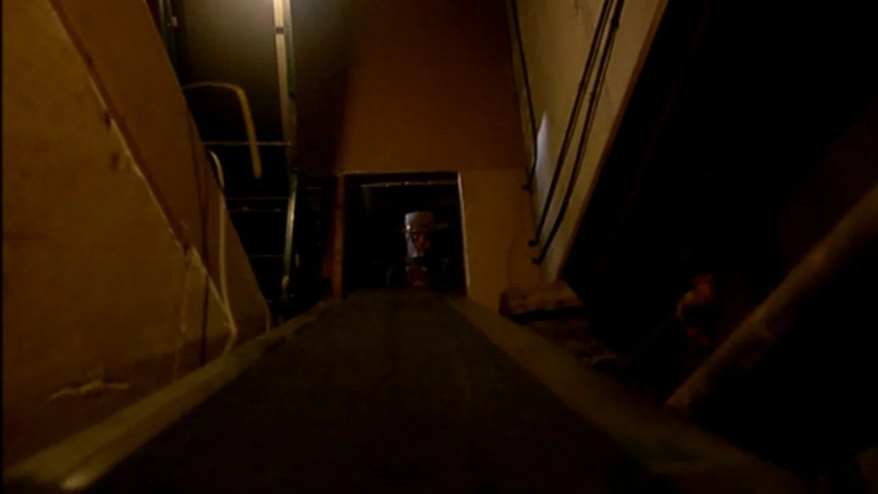 Murder Party movie still: wide — An industrial space features a long, dark conveyor belt leading into a lit doorway. A man wearing a hat stands at…