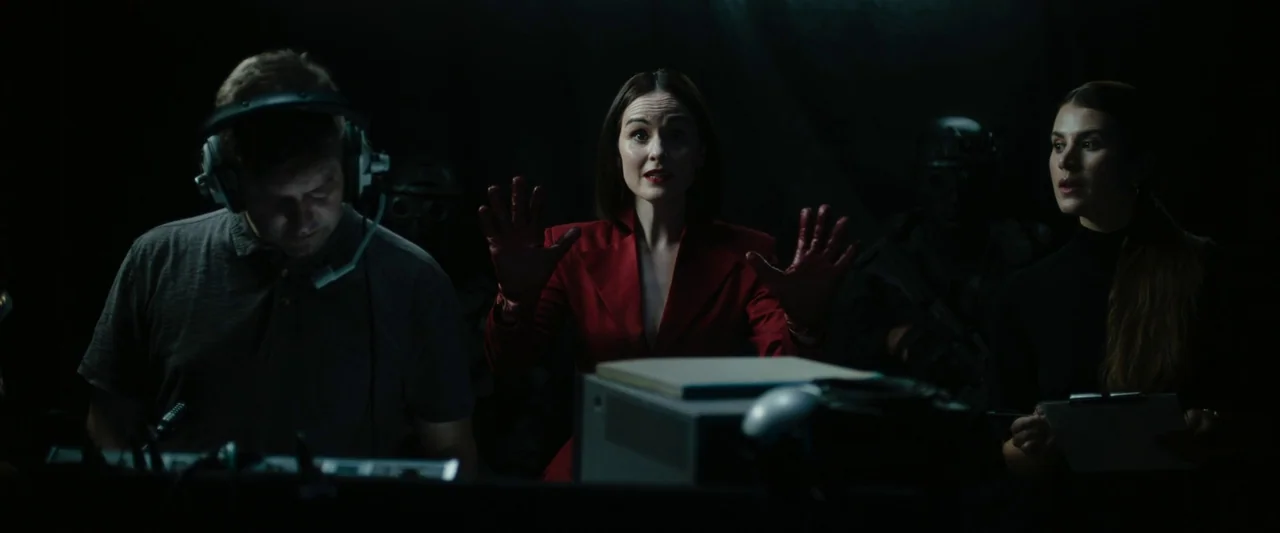 Boy Kills World (2023) movie still: medium — A dark, dimly lit technical room with figures obscured by low-key lighting. The central woman in a…